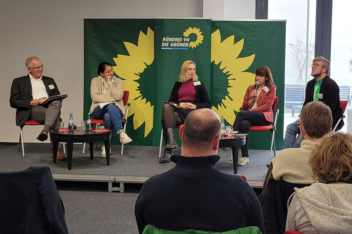 Diskussion beim Thementag gegen Rechts in Hannover. Eine leicht erhöhte Bühne, im Hintergrund 3 grüne Aufsteller im Grünen Parteidesign (dunkelngrün mi gelber Sonnenblume) Ddavor sitzen nebeneinander auf roten Stühlen (vlnr) Dirk Pejril, Filiz Polat, Greta Garlichs, Dr.in Julia Reuschenbach, Michael Lühmann. Es stehen 2 kleine schwarze runde Tische mit Wasserflaschen und Gläsern dabei. Im Vordergrund blickt man auf einige der Zuschauenden von hinten. Sie sitzen ebenfalls auf roten Stühlen.